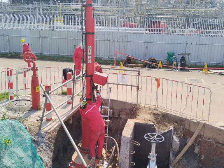 Workers in red safety gear perform a hot tapping procedure on an underground pipeline at a petrochemical plant; industrial process equipment and structural frameworks are visible in the background, with safety barriers and excavation site secured.