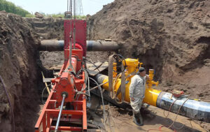 Red hot tapping equipment mounted on in-service natural gas pipeline (yellow fittings), with worker monitoring in excavation pit for hot tapping services