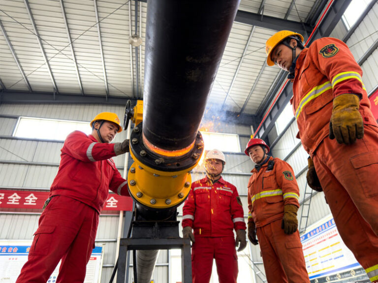Professional, wide-angle photo of a safe industrial worksite. A crew in full PPE (helmets, fire-resistant suits) is gathered around a pipeline where a yellow hot tap machine is installed. The scene is well-lit, with safety banners in the background. Photorealistic, high detail.