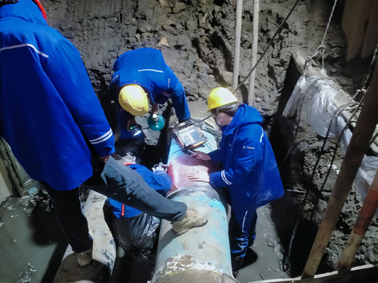 A team of workers in blue safety gear and yellow hard hats performs on-site pipeline repair in an excavated trench, using equipment to address pipeline damage.