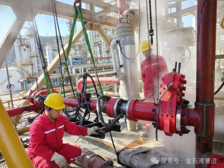 Two technicians in red uniforms and yellow safety helmets operating high-pressure hot tapping equipment on a red pipeline in an industrial facility with visible steam.