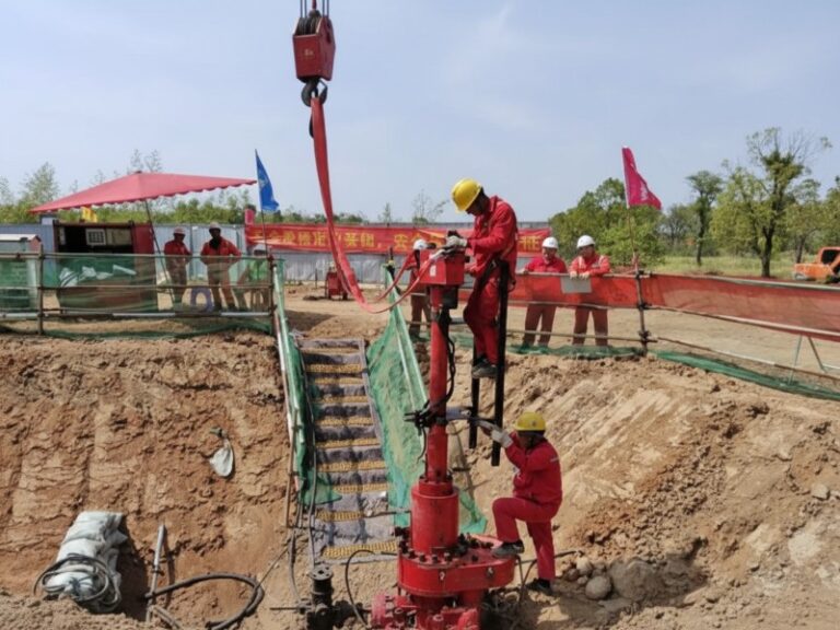 Industrial workers in red safety uniforms and hard hats operating a red JSW hot tapping machine for pipeline maintenance at an excavation site.