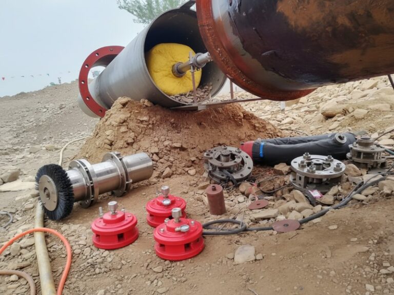 A collection of oil and gas pipeline maintenance equipment including red mechanical plugs, a pipeline cleaning brush tool, and line stop components laid out near a large steel pipe at a construction site.