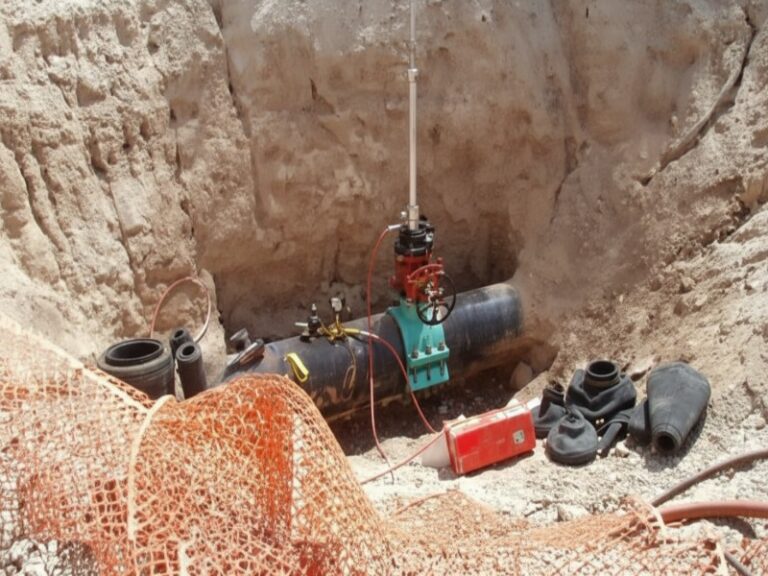 An industrial pipeline hot tapping operation in an open excavation pit, showing the specialized valve and tapping machine assembly on a large black pipe.
