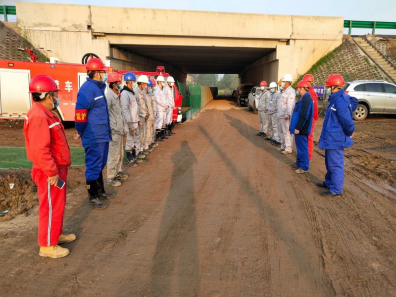 Construction workers in safety gear and helmets gather for a safety briefing at the Anhe and Anhao refined oil pipeline relocation project site in Shucheng Hangbu Town, including fire safety equipment in the background.