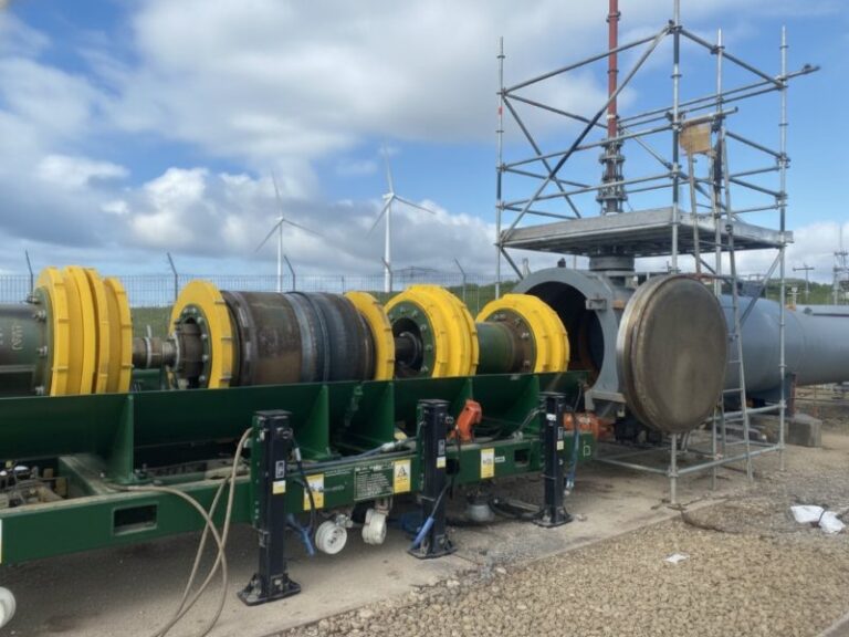 An innovative pipeline isolation tool with yellow discs and a heavy-duty metallic body is positioned on a green loading frame in front of a large pipeline opening, with wind turbines visible in the distance under a cloudy sky.