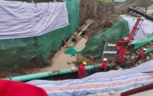 Industrial construction crew performing oil pipeline hot tapping, plugging, and connection services in an excavated trench. The site shows workers in red safety suits near large pipelines and machinery, with protective tarps covering the surrounding soil.