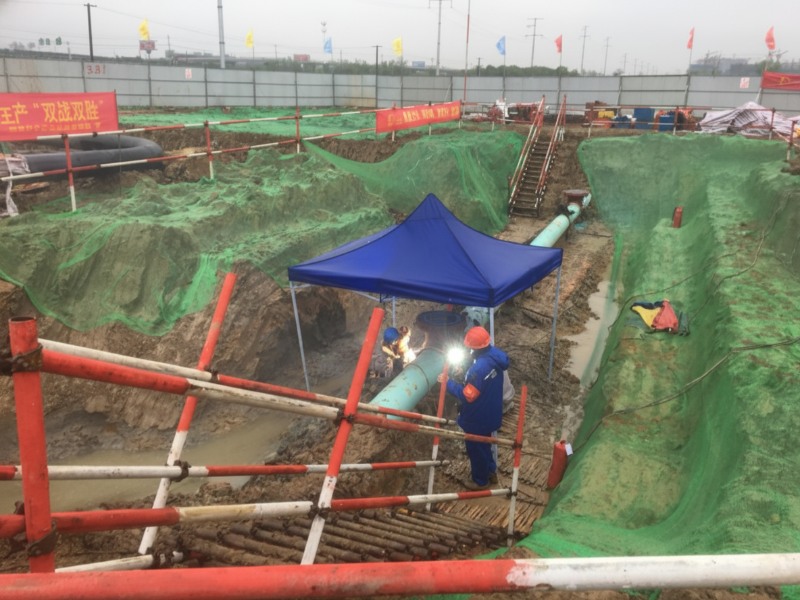 Workers performing welding and oil recovery on the Anhe and Anbo product oil pipeline relocation project in Shucheng Hangbu Town, showing an excavated construction pit with green safety netting and a protective blue canopy over the pipeline joint.