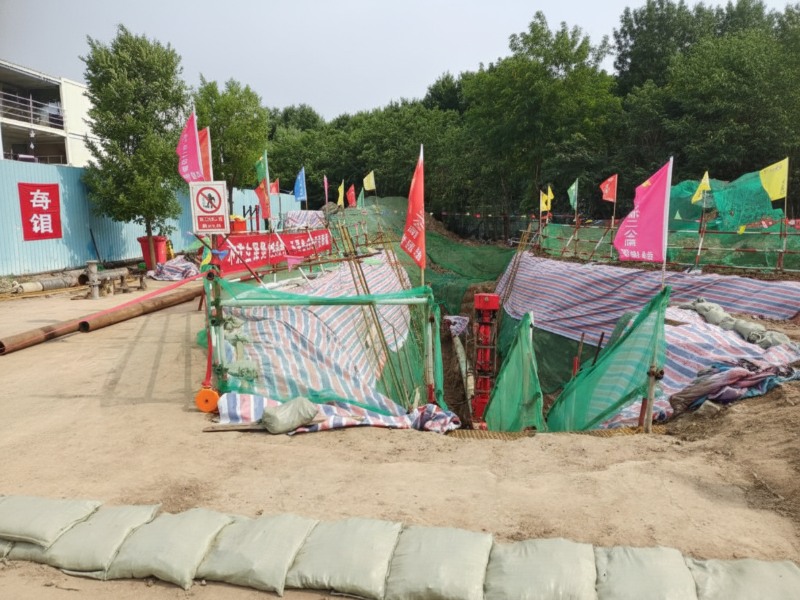 A construction site for pipeline replacement and sealing featuring an excavation pit protected by green safety netting and striped tarps with red warning flags and sandbags in the foreground.