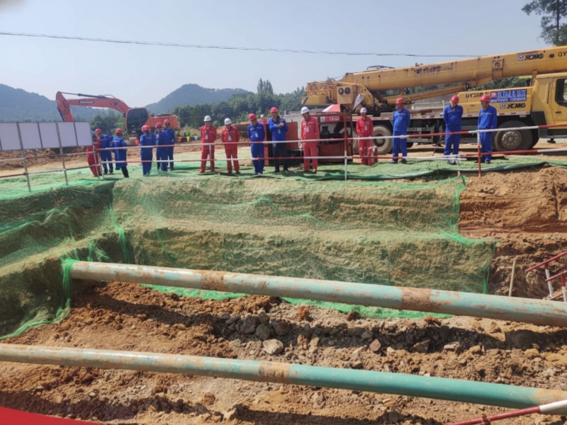 A group of engineers and workers at a pipeline relocation and hot tapping construction site in Anhui, China, with heavy machinery and large industrial pipes in view.