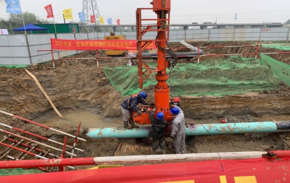Construction workers in safety gear operating a large orange hot tapping machine on a pipeline at the Anhe-Anhao refined oil pipeline relocation and oil recovery site in Shucheng, China.