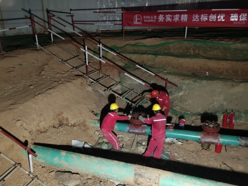Workers in red safety uniforms and yellow hard hats performing hot tapping and plugging on a high-pressure green oil pipeline at night during the Anhe and Anhao pipeline relocation project in Anhui.