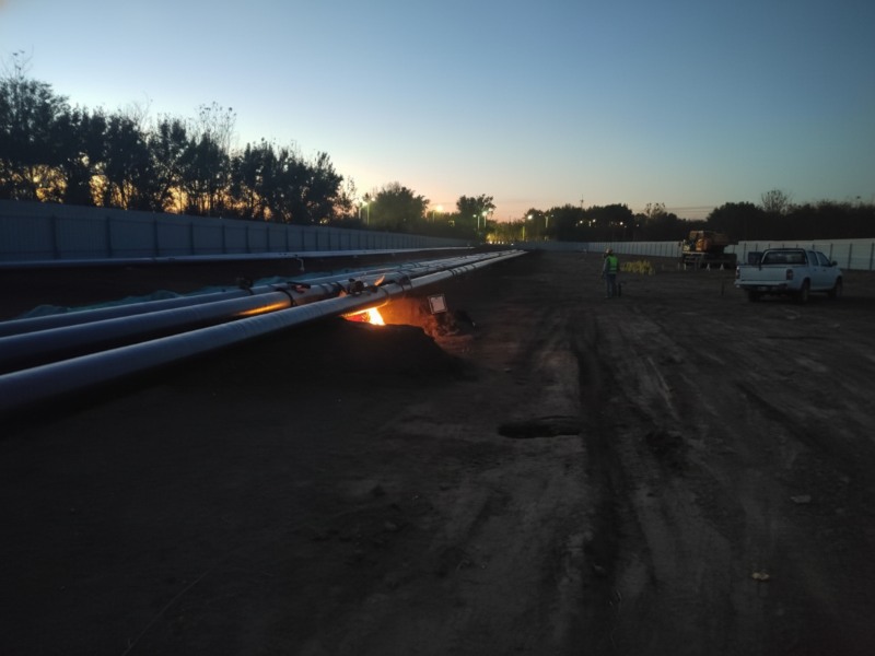 Construction site of the Wenchao Jianhe pipeline relocation project, showing large diameter Sinopec refined oil and aviation fuel pipelines laid out at twilight with a bright welding light visible beneath the pipes.