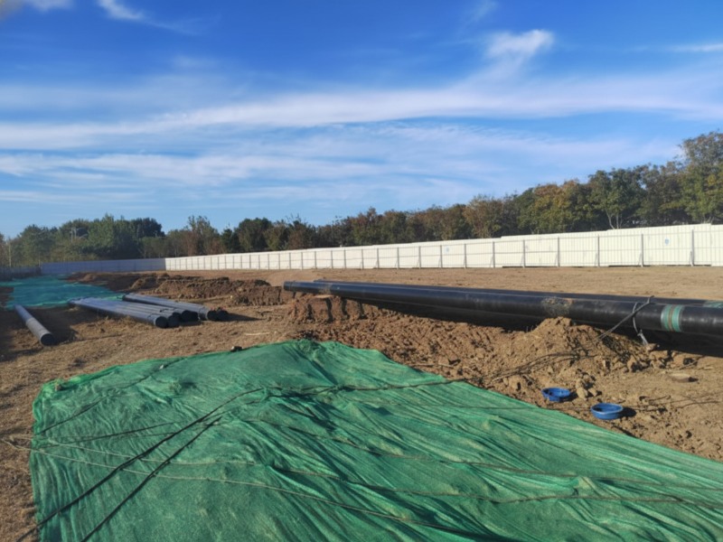 An industrial construction site for the Wenchao Relief River pipeline relocation project, featuring large black steel pipes for oil and fuel transport laid out on a dirt field with green safety netting and a white perimeter fence.