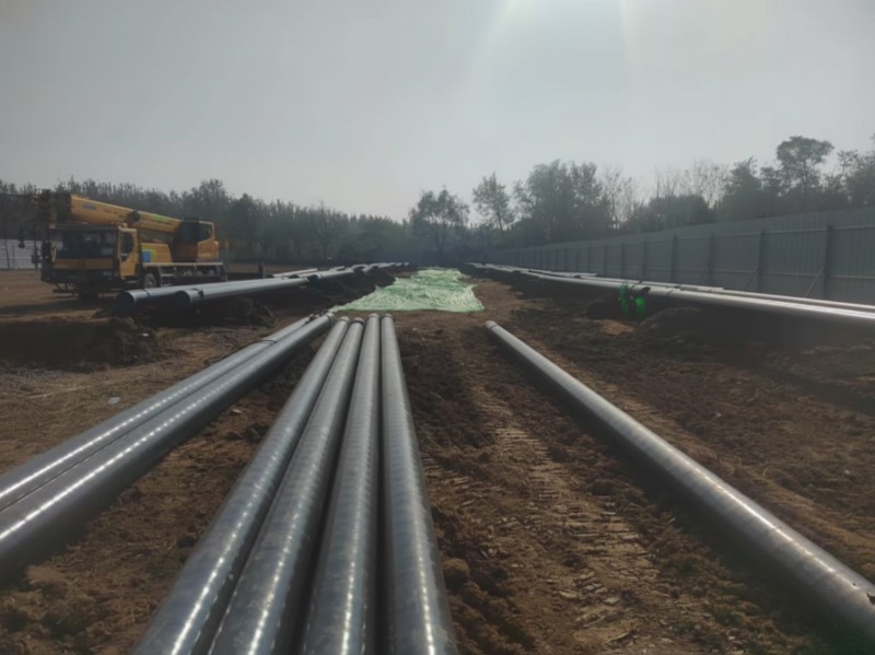 Large industrial oil pipelines laid out for relocation at the Wenchaojian River engineering site with a yellow construction crane in the background.