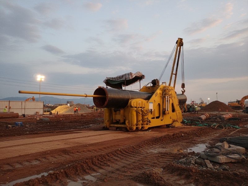 A large yellow industrial pipe bending machine performing a pipeline cold bending service on a construction site with workers and heavy equipment in the backgound