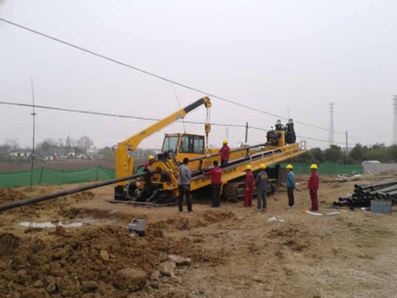 A large yellow crawler-mounted horizontal directional drilling (HDD) rig being operated by workers in safety gear on a construction site for underground pipe installation.