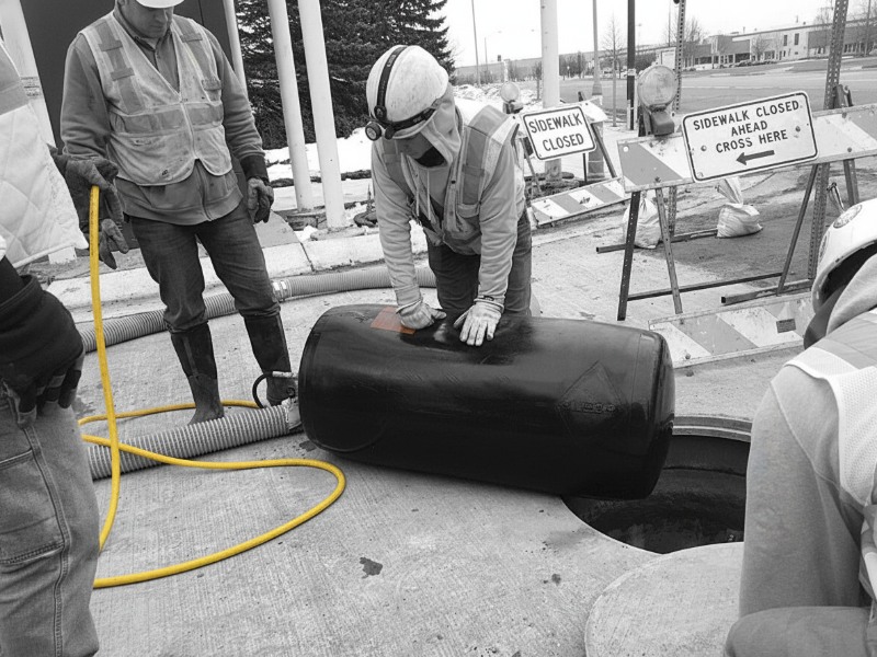 Construction workers in safety gear preparing a large black inflatable pipe plug for sewer pressure testing near an open manhole.