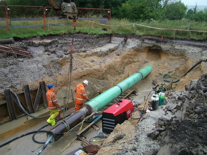 Two construction workers in high-visibility safety gear overseeing the pipe ramming installation of a large green steel casing pipe in a muddy trench.