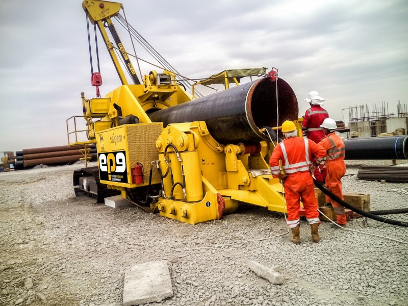 Industrial workers in orange safety gear monitoring a large yellow machine during a professional pipeline cold bending service at a construction site