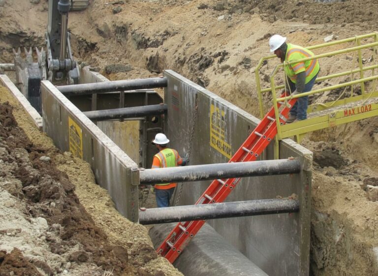 Construction workers using a metal trench shield system for safety while working in a deep open excavation.