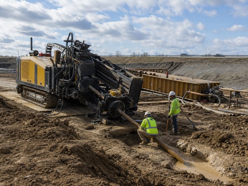 Large horizontal directional drilling rig working beside a highway crossing with drill pipe entering the ground at a shallow angle