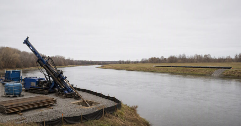 Horizontal directional drilling rig on a crossing site with a curved bore path under a river, showing the rig pad, drill pipe and staged product pipe string
