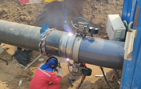 An automated orbital welding machine precisely joining sections of a large diameter industrial pipeline, monitored by a professional welder at a construction site.