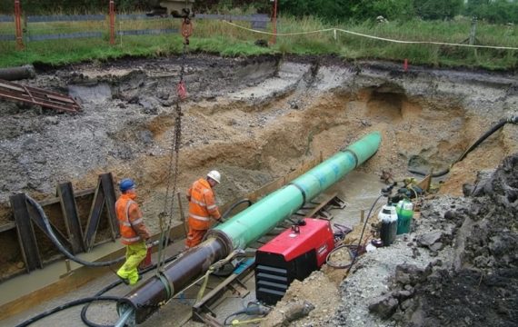 Two construction workers in high-visibility safety gear overseeing the pipe ramming installation of a large green steel casing pipe in a muddy trench.